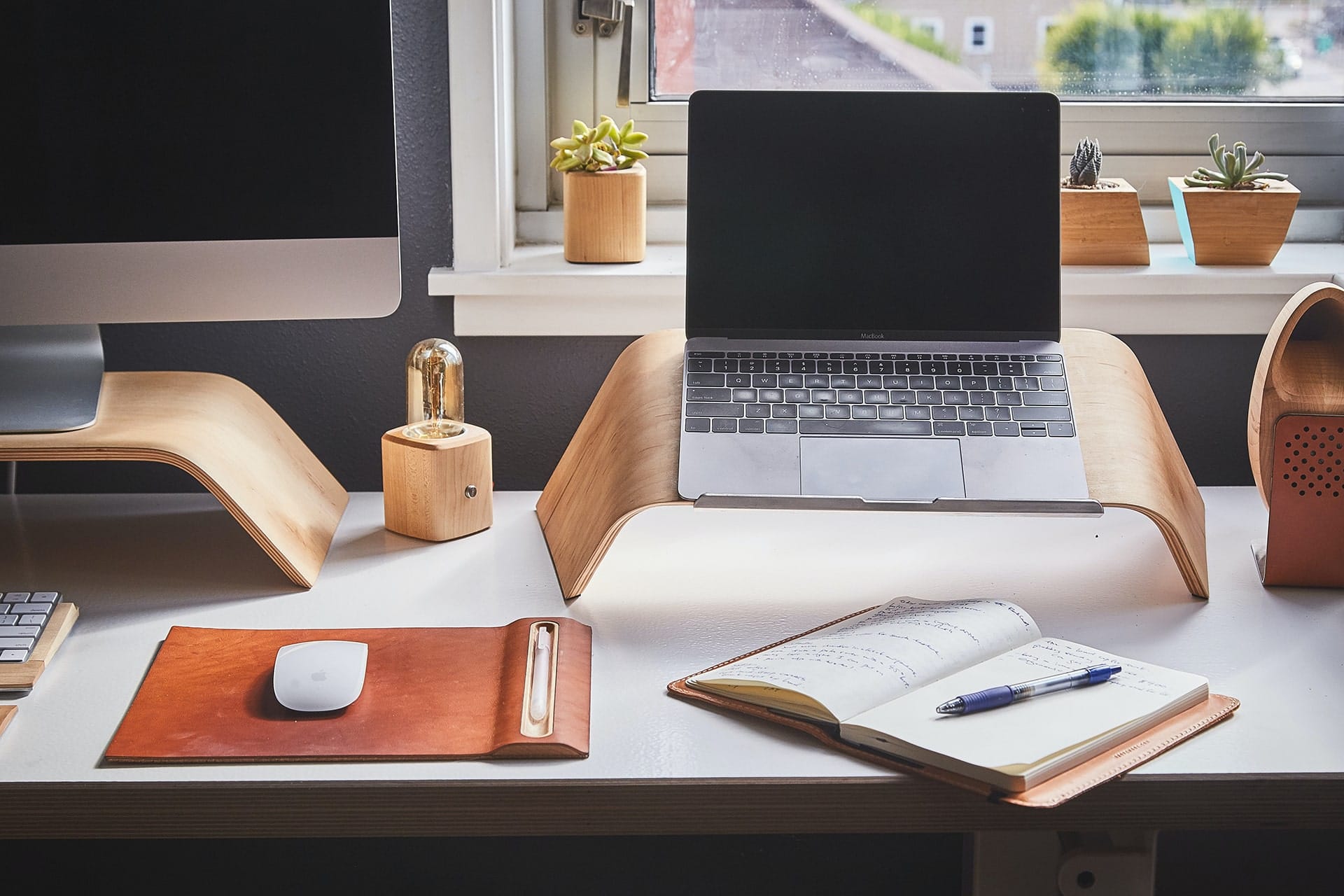 Tidy desk with a laptop on a wooden stand, open notebook and pen, beside a sunlit window