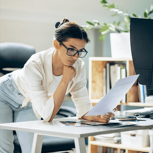 Person leaning over a desk reviewing a printed document beside a monitor