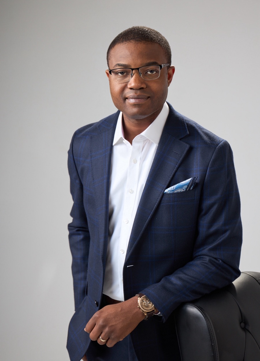 Abraham Oni, Managing Principal of Innovating Chaos, standing beside a leather chair in a navy checked suit against a light grey studio backdrop.