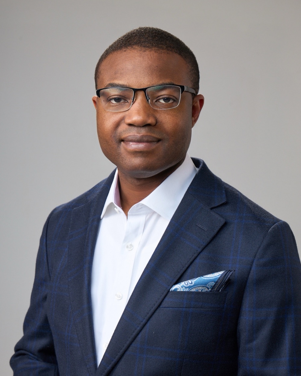 Abraham Oni, Managing Principal of Innovating Chaos, in a navy checked suit against a light grey studio backdrop.